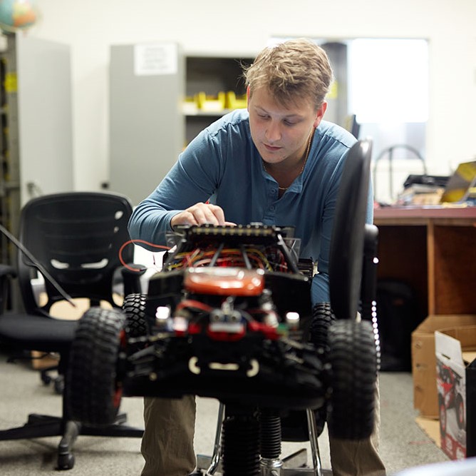 A male student sits behind a small electronic car with wires leading to a display he is holding.